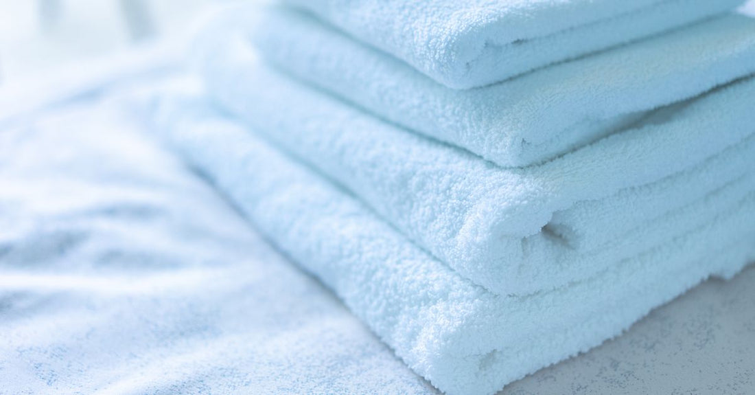 Stack of soft cotton facial towelettes and a rolled microfibre cloth on a light bathroom surface, showing textures for how to choose cloths & towelettes.
