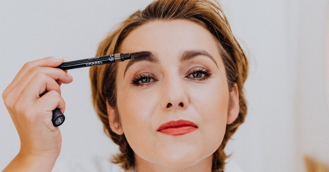 A selection of eyebrow pencils, powders and gels in various shades swatched on a neutral background, illustrating an eyebrow color buying guide.