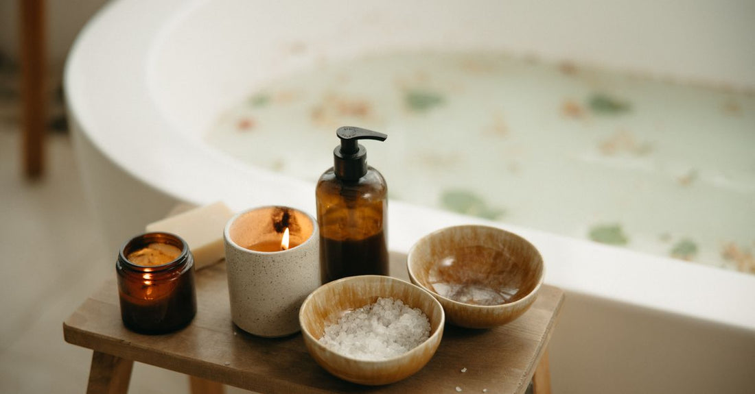 A flatlay of bath and bathing accessories including a bottle of bath oil, a jar of bath salts, and a wooden bath brush on a marble surface.