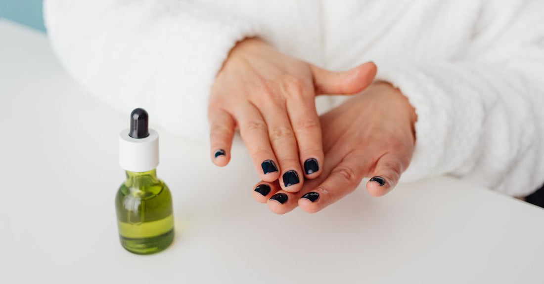How to use cuticle oil properly: a glass dropper bottle of nourishing cuticle oil surrounded by fresh botanicals on a light marble surface.