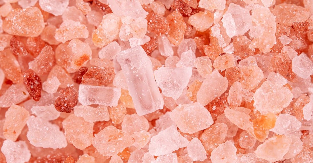 A clear glass jar of mineral-rich bath salts with a scoop, on a bright summer bathroom shelf.