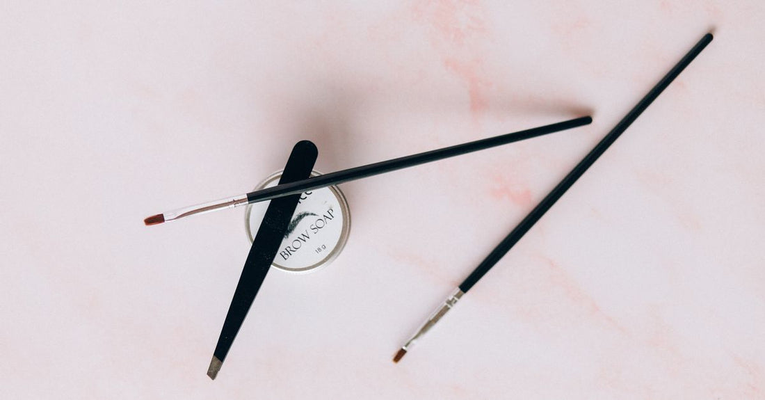 A flatlay of stainless steel eyebrow grooming tools including tweezers, scissors, and a spoolie brush on a white background, demonstrating how to groom eyebrows at home.