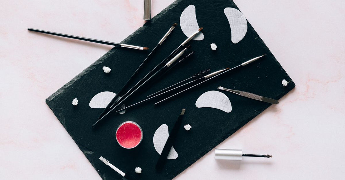 A flatlay of eyebrow grooming tools: precision scissors, tweezers, an angled brush, and brow spoolie on a light marble surface, illustrating how to shape eyebrows at home.
